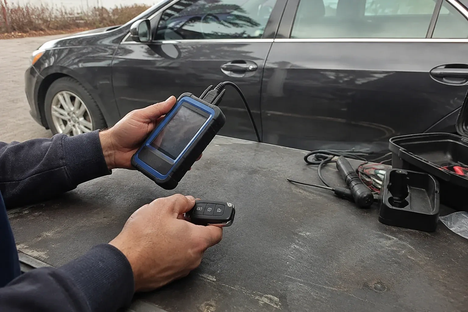 Hands holding a blue-edged electronic device and car-key fob at a worktable beside a parked car. — Transponder Key Programming, Oro-Medonte