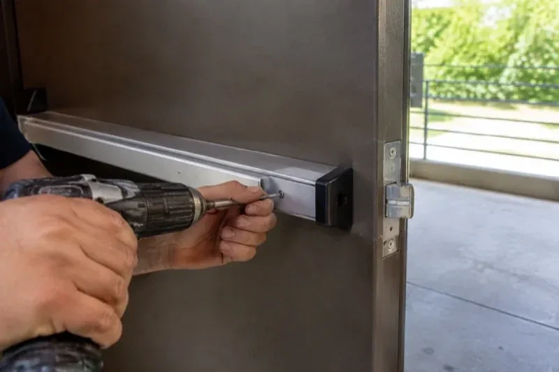 Hands using a power driver at the end of a horizontal push bar on an open metal door. — Panic & Exit Hardware, Oro-Medonte